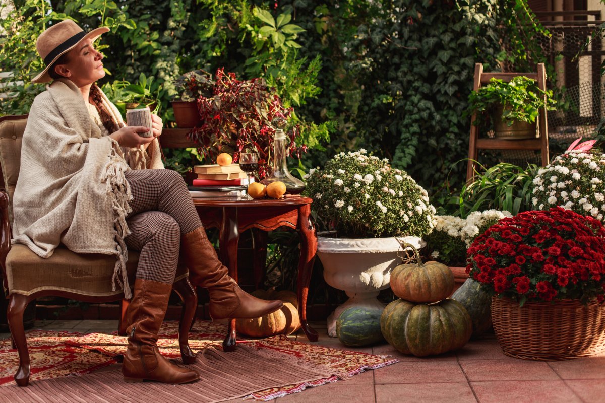 Woman wearing a blanket draped over her and sitting outside with area rug under her feet and pumpkin decor next to her.