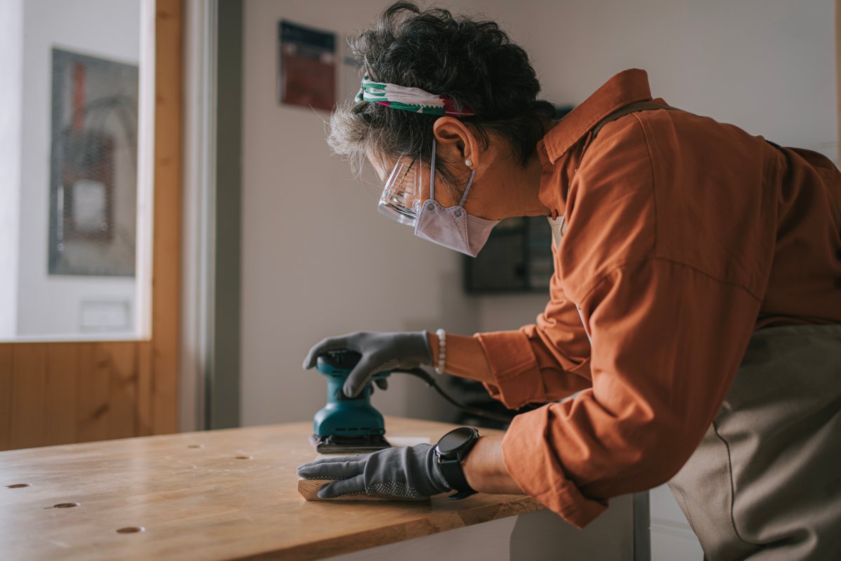 Woman using sander while working on a piece of wood in woodshop and wearing gloves, a mask, and protective glasses.