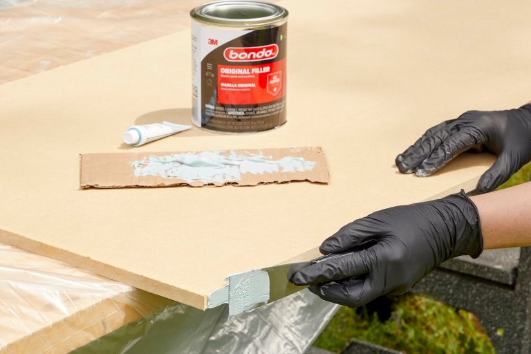 Hands using a putty knife to spread Bondo original filler on edge of MDF panel.