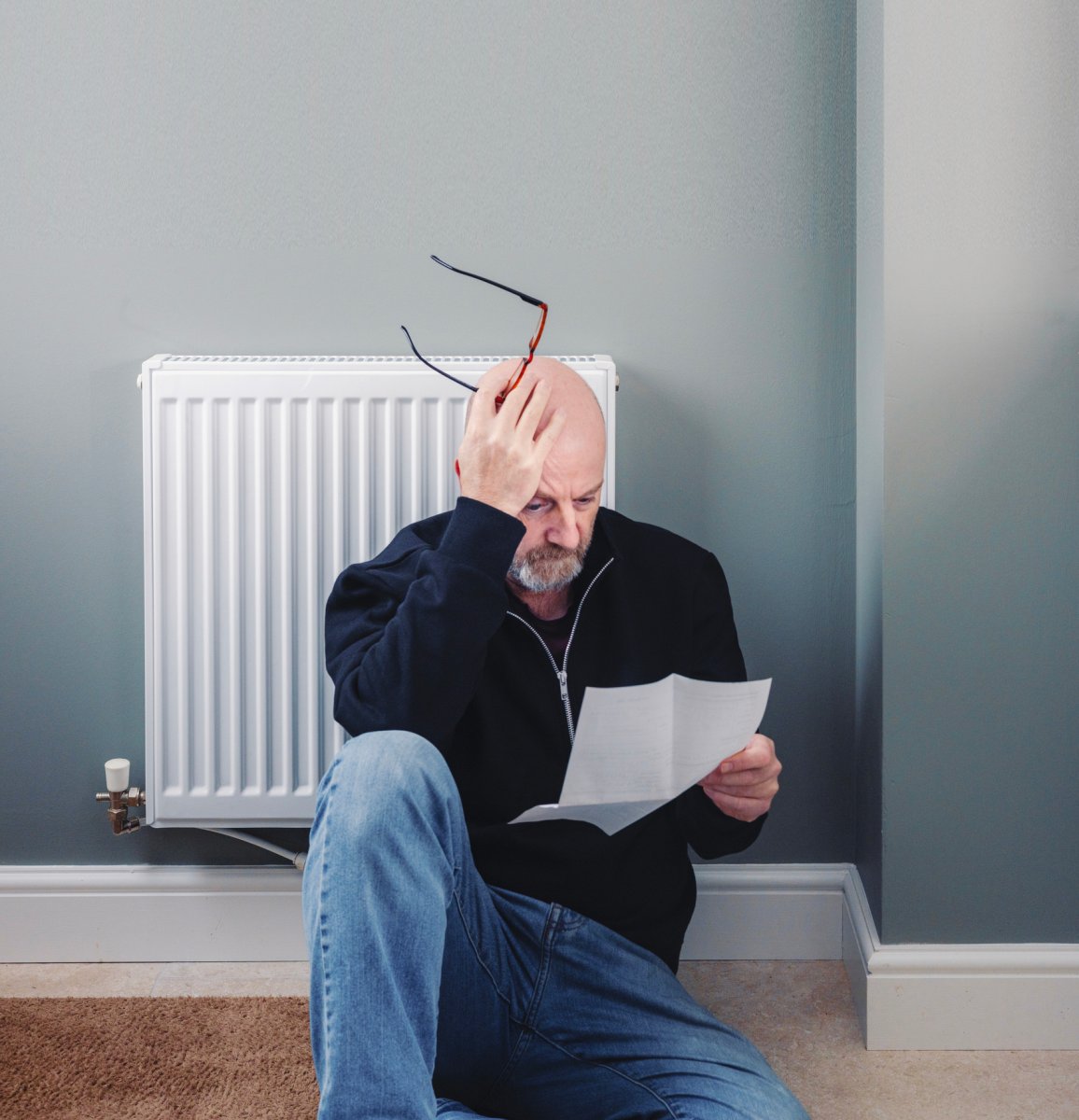 Worried man looking at his heating bill sitting down leaning against a radiator with his hand on his head