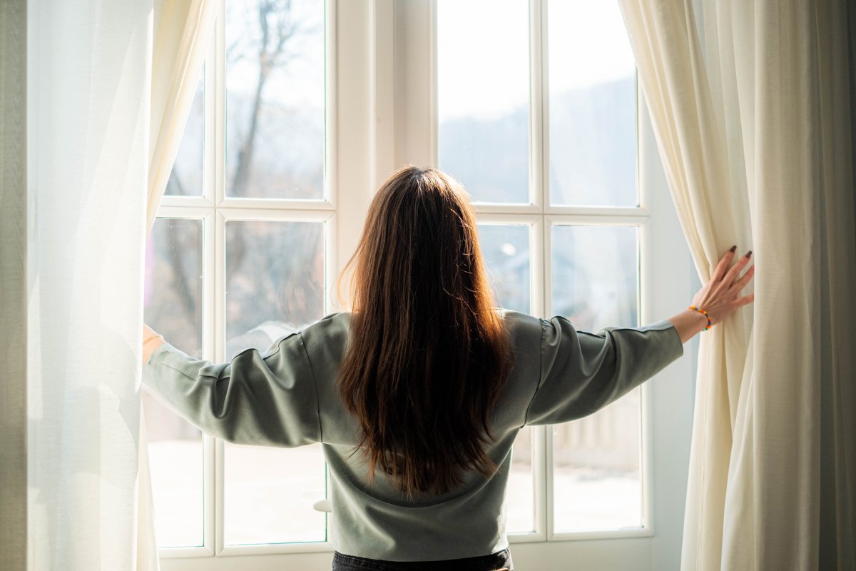 A woman pulling back the curtains so that the sun can enter the room.