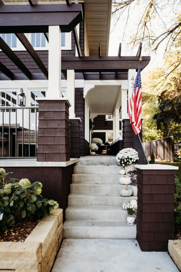 A brown, coastal home with an American flag and potted plants on the front porch.