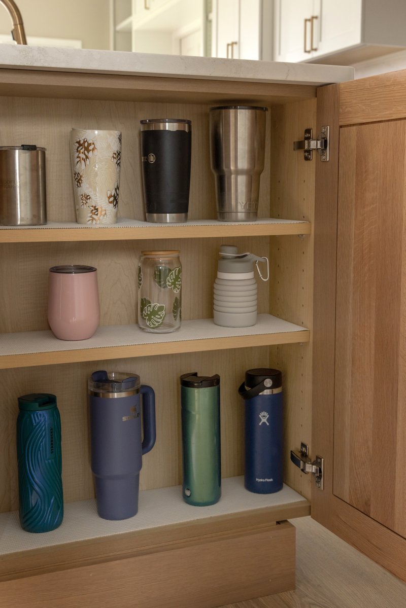 A shallow kitchen cabinet for storing water bottles and to-go coffee mugs.
