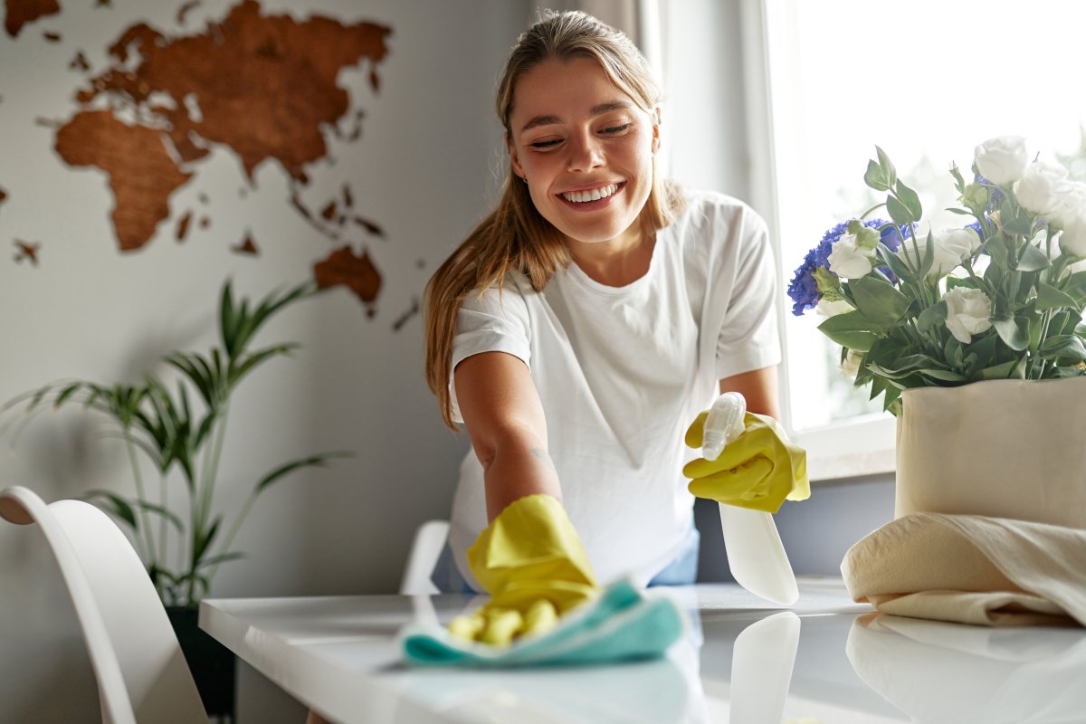 A cheerful young woman in bright yellow gloves cleaning a table in a room in her house.