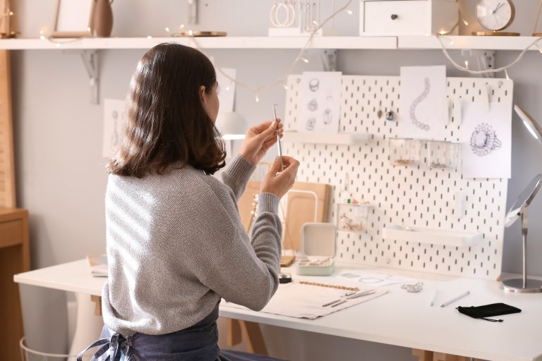 Woman designing jewelry in an art studio with a white pegboard displaying items.