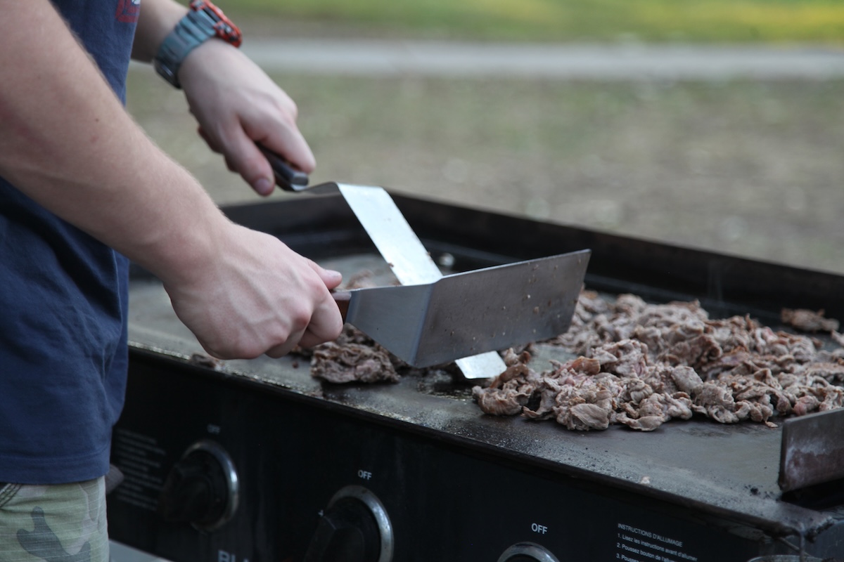 Person wearing a blue T shirt uses spatula to cook chopped meat on a flat-top griddle.