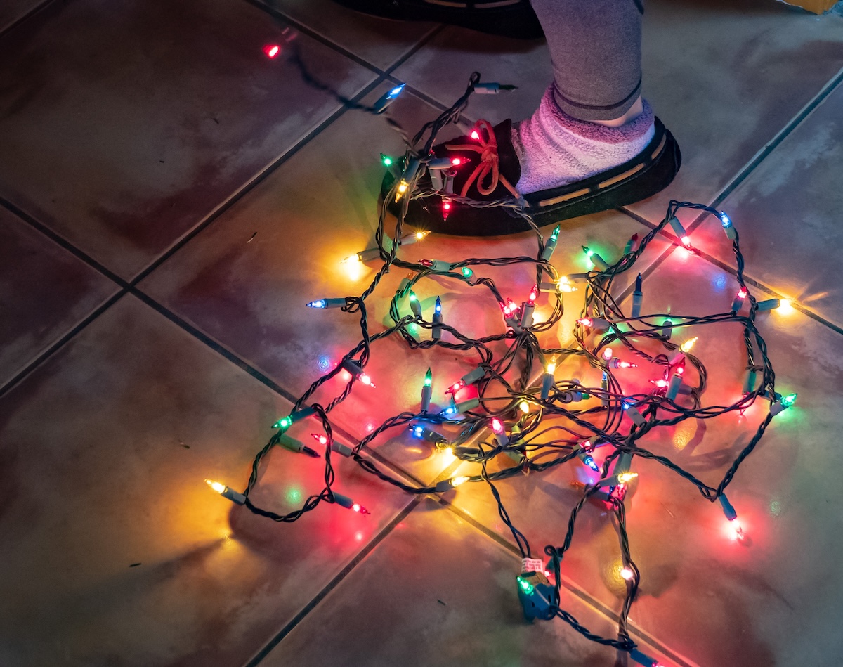 Tangled, lit, multicolored Christmas lights around the feet of a person wearing slippers.