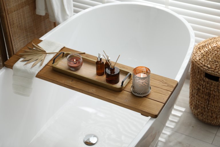 Wooden tray with cosmetic products, burning candles, reed air freshener and towel on bath tub in bathroom