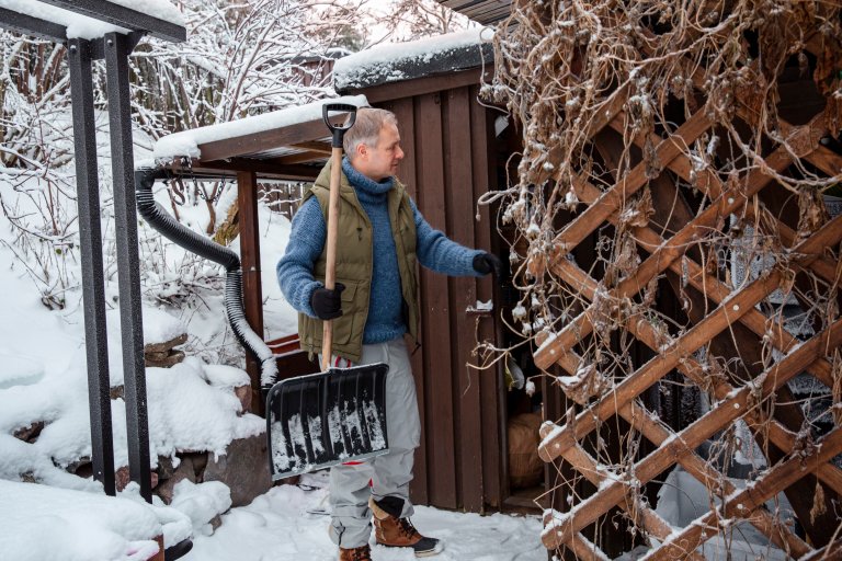 Middle-aged man taking a shovel from shed and heading into snowy yard, performing seasonal winter chores, outdoor lifestyle in cold weather.