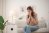 Young woman sits on a couch, sneezing and holding a tissue to her nose while an air purifier operates nearby.