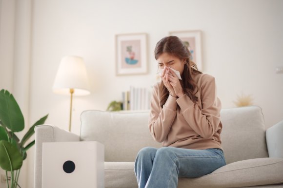 Young woman sits on a couch, sneezing and holding a tissue to her nose while an air purifier operates nearby.