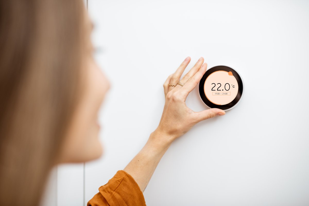 Woman regulating heating temperature with a modern wireless thermostat installed on the white wall at home.