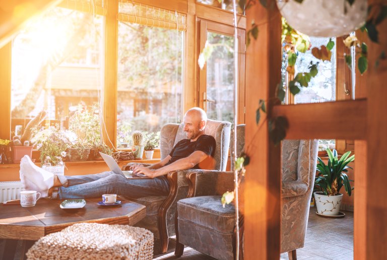 Man dressed black T-shirt and jeans sitting in a comfortable armchair, using a modern slim laptop and drinking tea in house sunroom.
