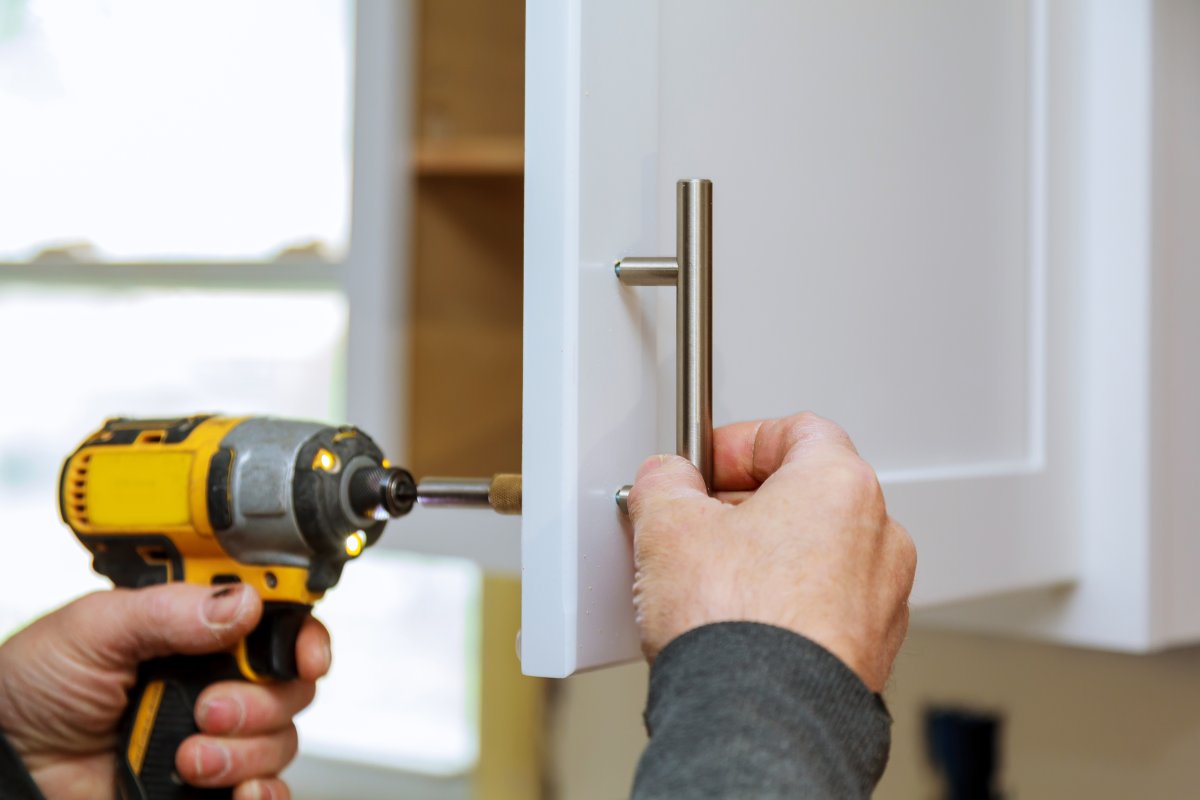 A man sets a new handle on a white kitchen cabinet with an electric screwdriver.