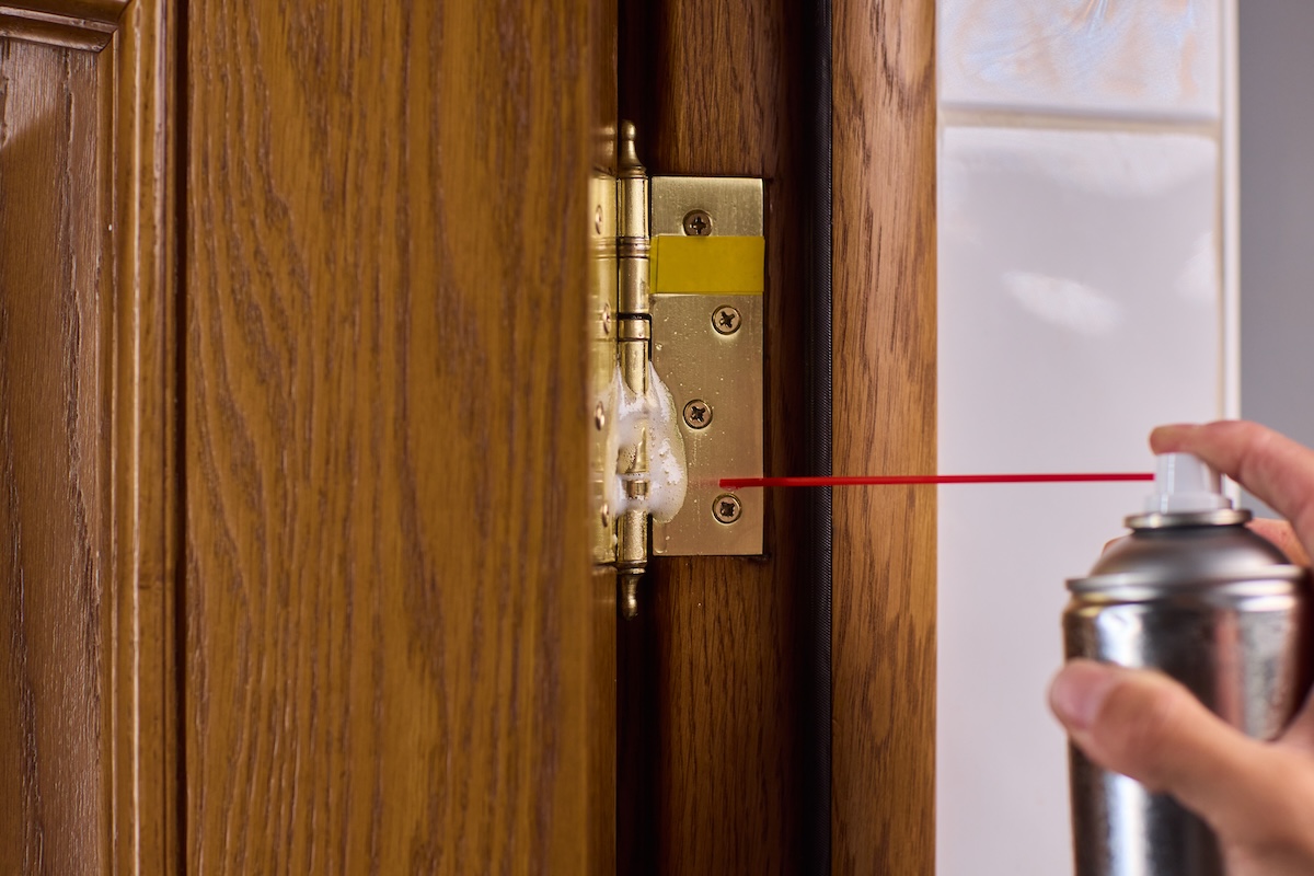 A hand spraying lubricant on door hinge to eliminate squeaks.
