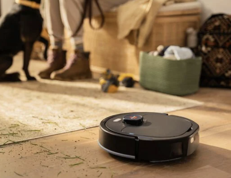 BISSELL FlexClean™ FurForce™ 2-in-1 Vac and Mop Robot in the foreground with a dog standing on the floor and a woman sitting on the couch in the background.