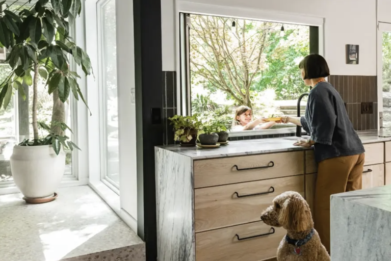 Woman handing a plate to a young girl through a large pass through window in her house.