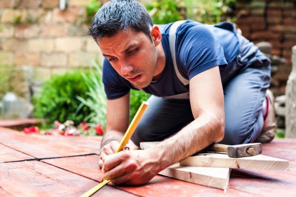 Young man uses pencil to measure something on the floor of a deck in deck construction/repair process.