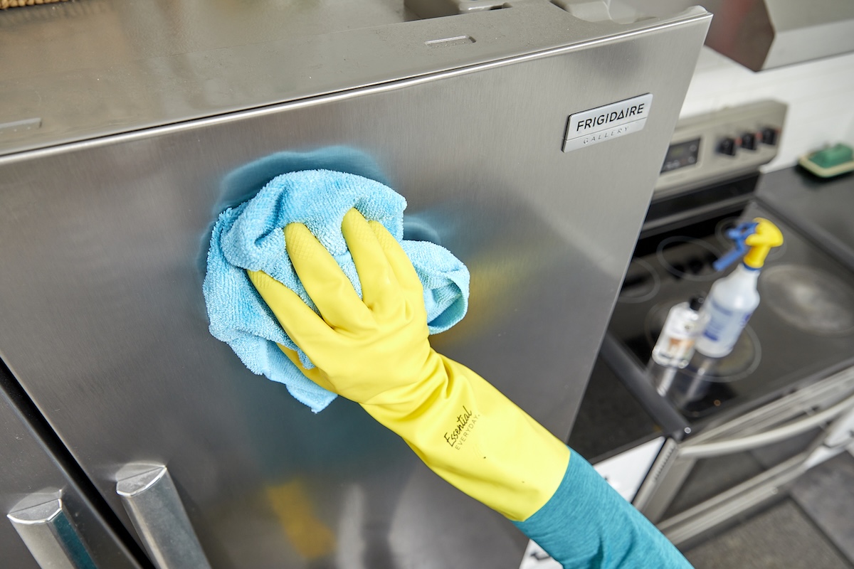 Person wearing yellow rubber gloves uses microfiber cloth to wipe a stainless steel refrigerator.