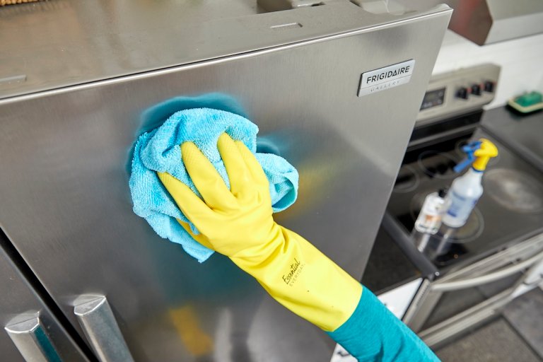 Person wearing yellow rubber gloves uses microfiber cloth to wipe a stainless steel refrigerator.