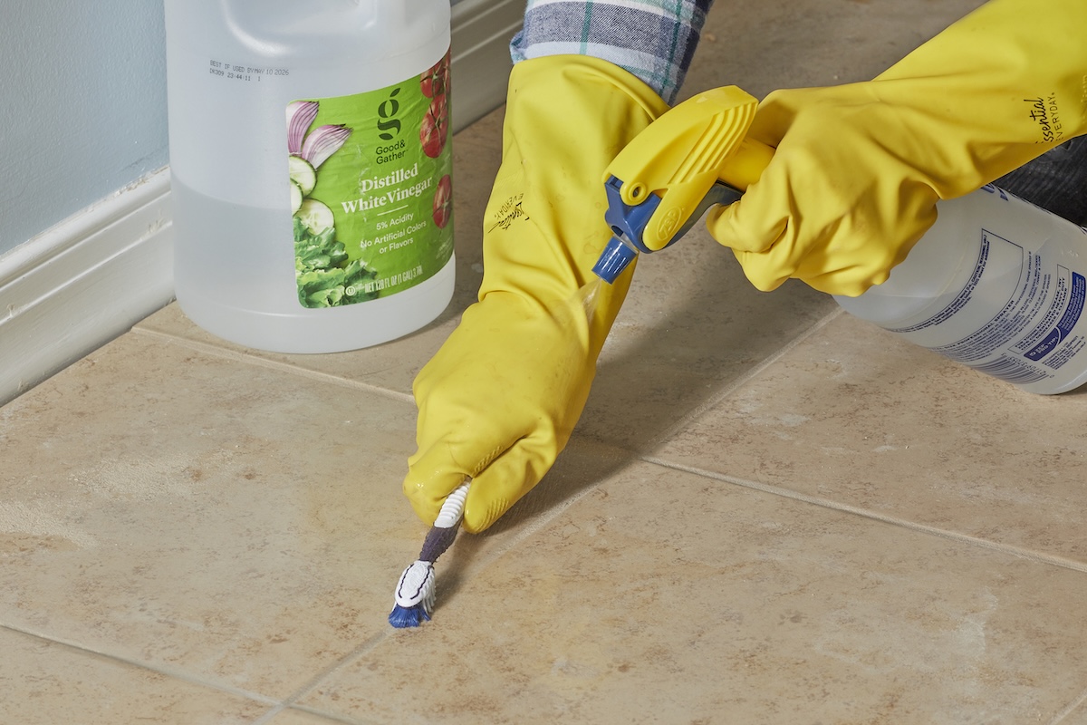Woman wearing rubber gloves sprays tile grout with spray bottle, and cleans it with small scrub brush, a large bottle of vinegar in the background.