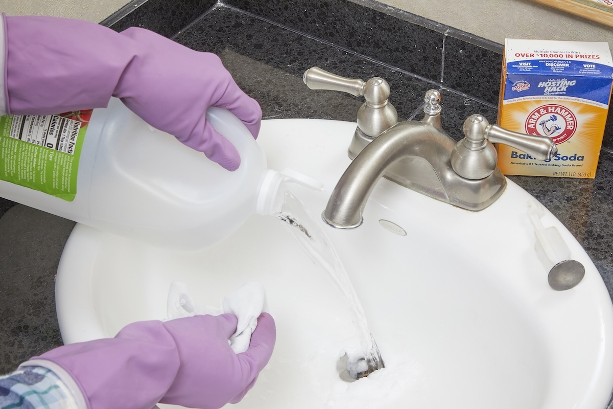 Woman wearing rubber gloves pours vinegar on top of baking soda into bathroom sink.