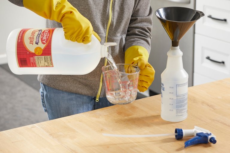 Woman pours vinegar from a jug into a measuring cup; a funnel sits in a spray bottle nearby.