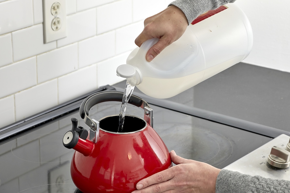 Woman pours vinegar from a gallon container into a red kettle on top of a glass cooktop.