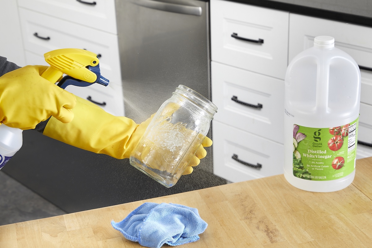 Woman wearing yellow rubber gloves sprays a mason jar with spray; a jug of vinegar is nearby on the counter.