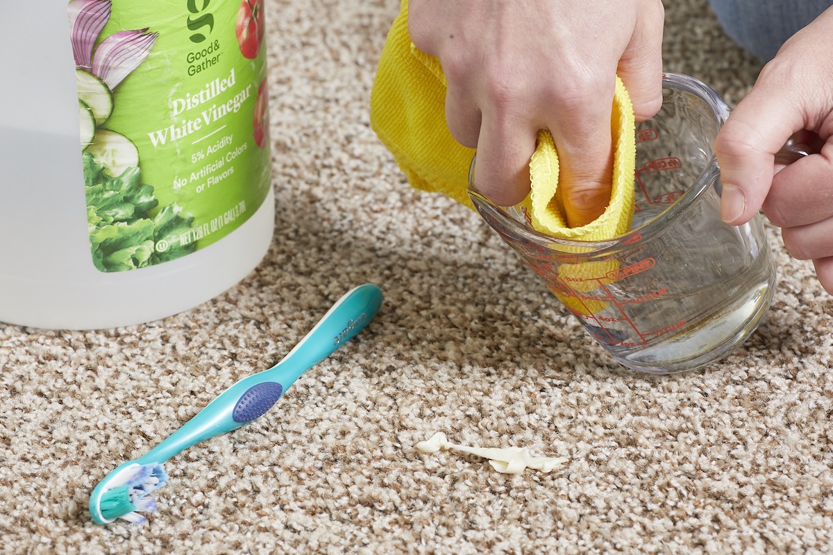 Woman dips yellow microfiber cloth into measuring cup with liquid, poised over brown carpeting. A toothbrush and jug of vinegar rest on the carpet nearby.