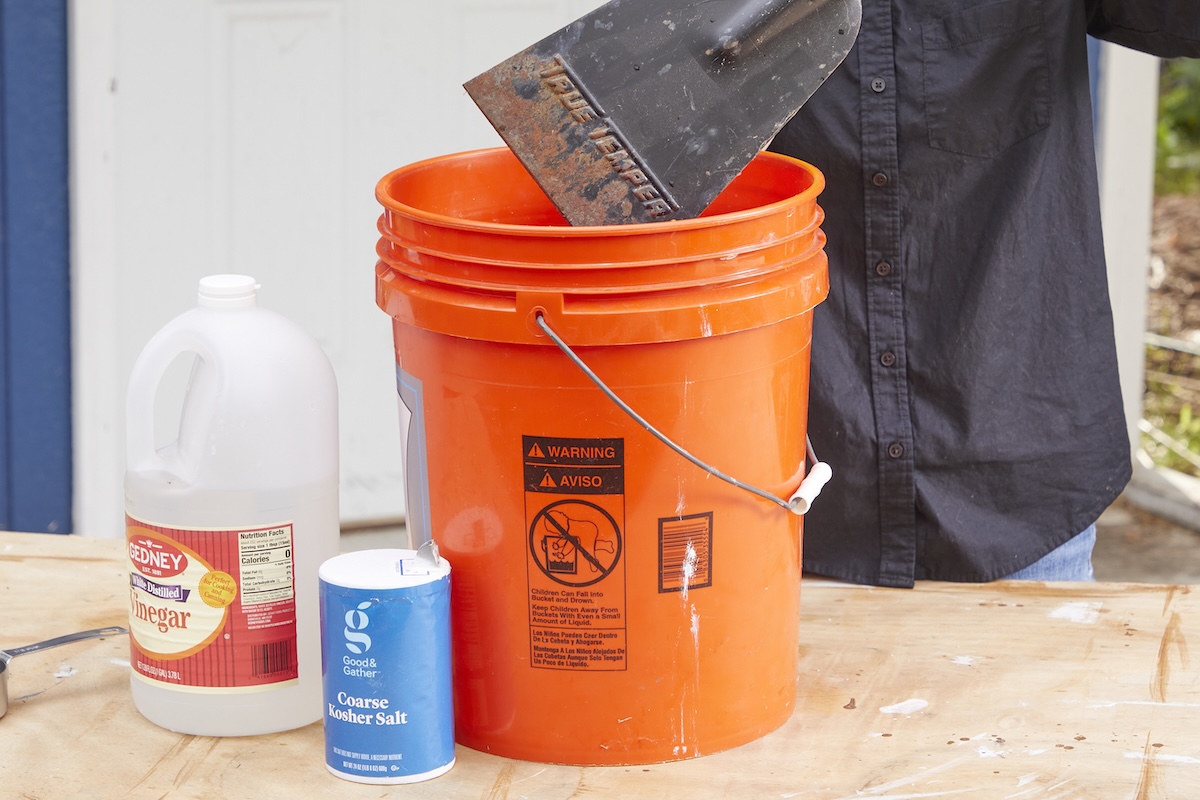 Woman dunks rusty shovel head into an orange 5-gallon bucket, containers of salt and vinegar nearby.