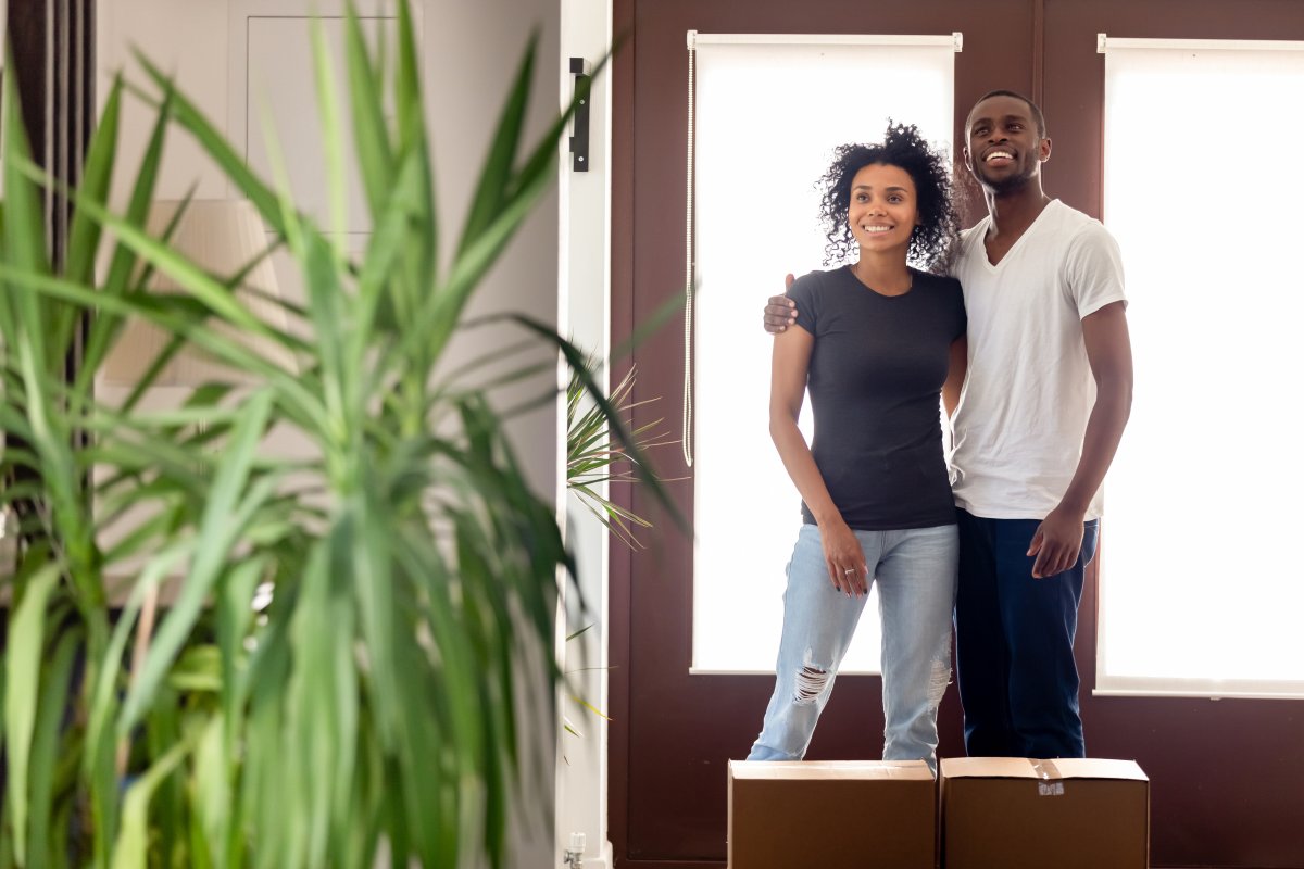 Smiling excited Black man and woman standing in entry of their new home with cardboard boxes.
