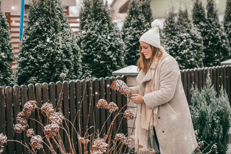 Young woman in winter clothes and knitted white hat with scarf in the winter garden with snow-covered plants.