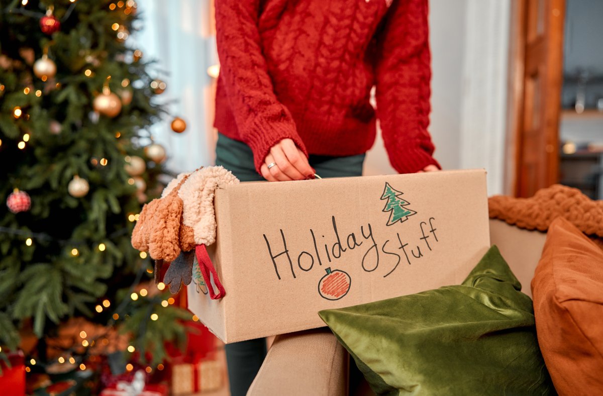 A woman closing a cardboard box labeled "Holiday Stuff" to store and a Christmas tree in the background.