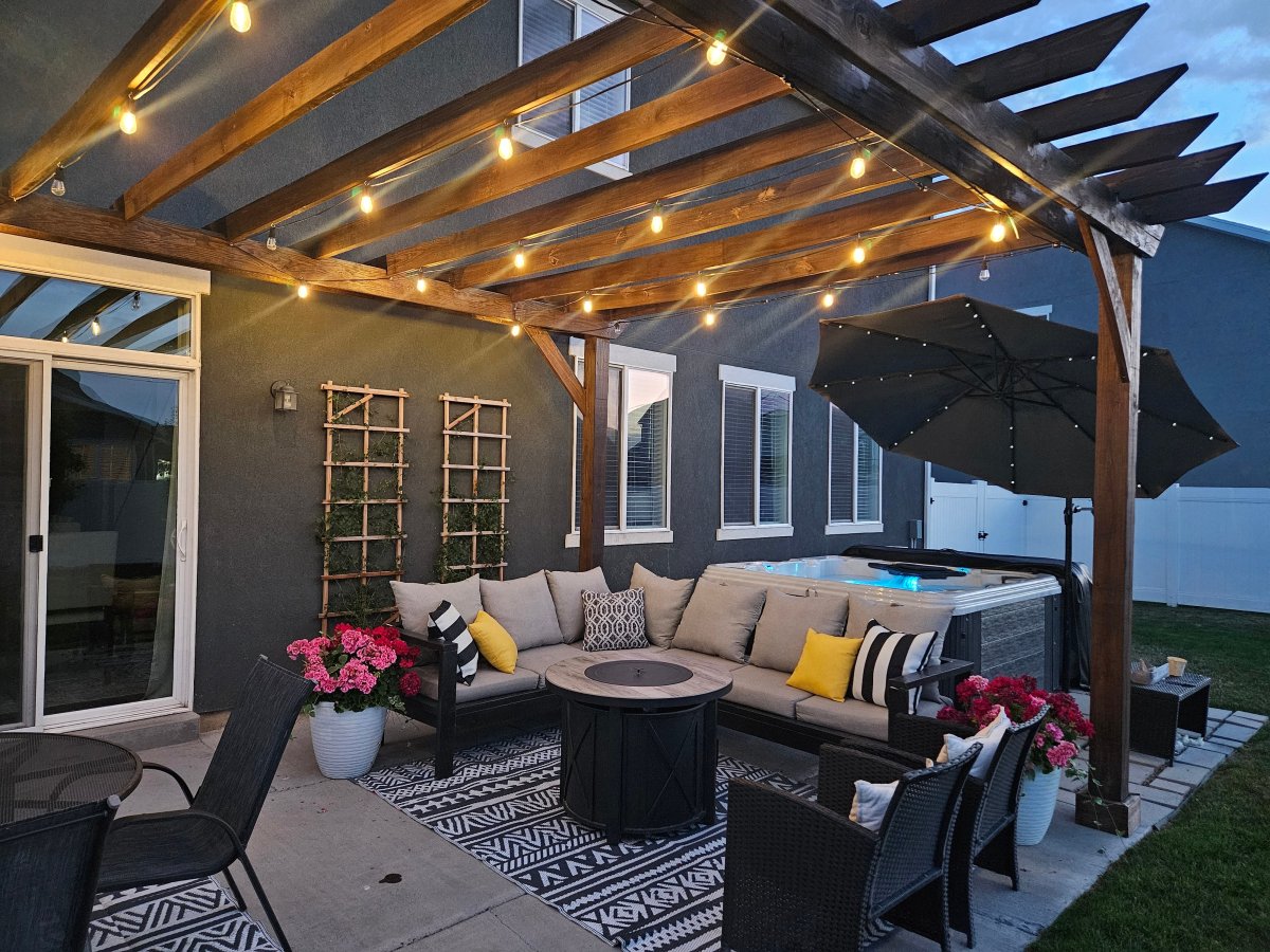Lit up pergola and outdoor patio with a grey L-shaped couch, striped and yellow pillows, black and white rugs, potted flowers, hot tub, fire pit.