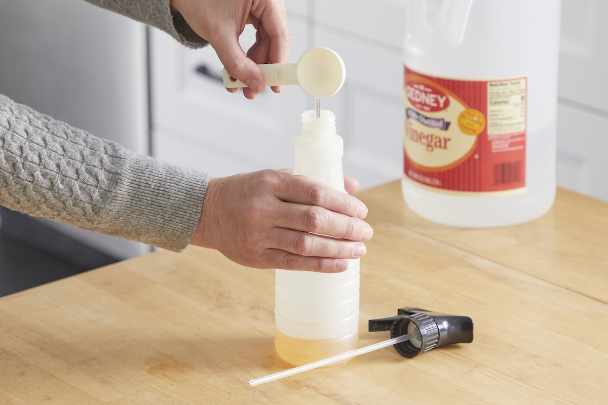 Woman uses a tablespoon to measure something into a spray bottle; bottle of vinegar in the background.