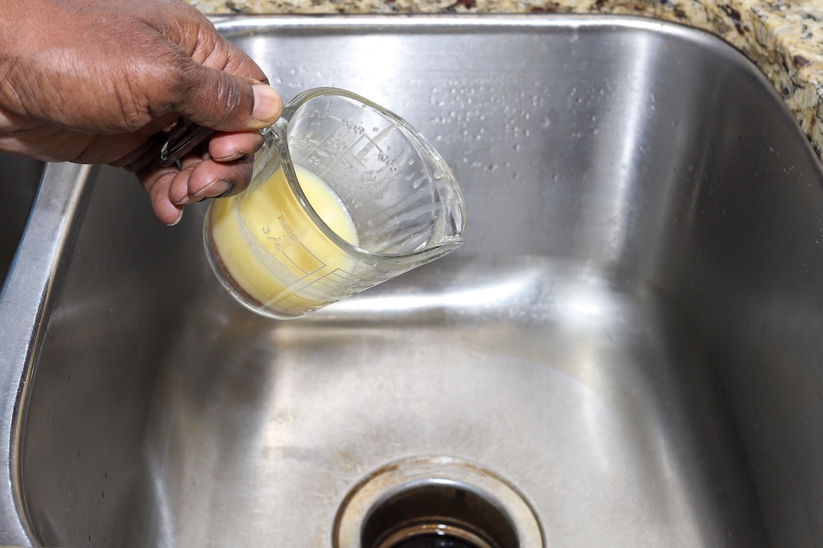 A person pouring grease from a measuring cup in a kitchen sink.