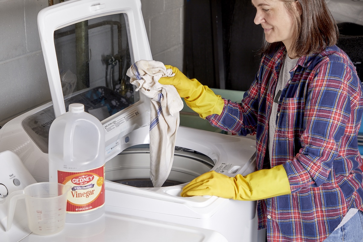 Woman tosses dish towels into a top-load washing machine, a jug of vinegar nearby.