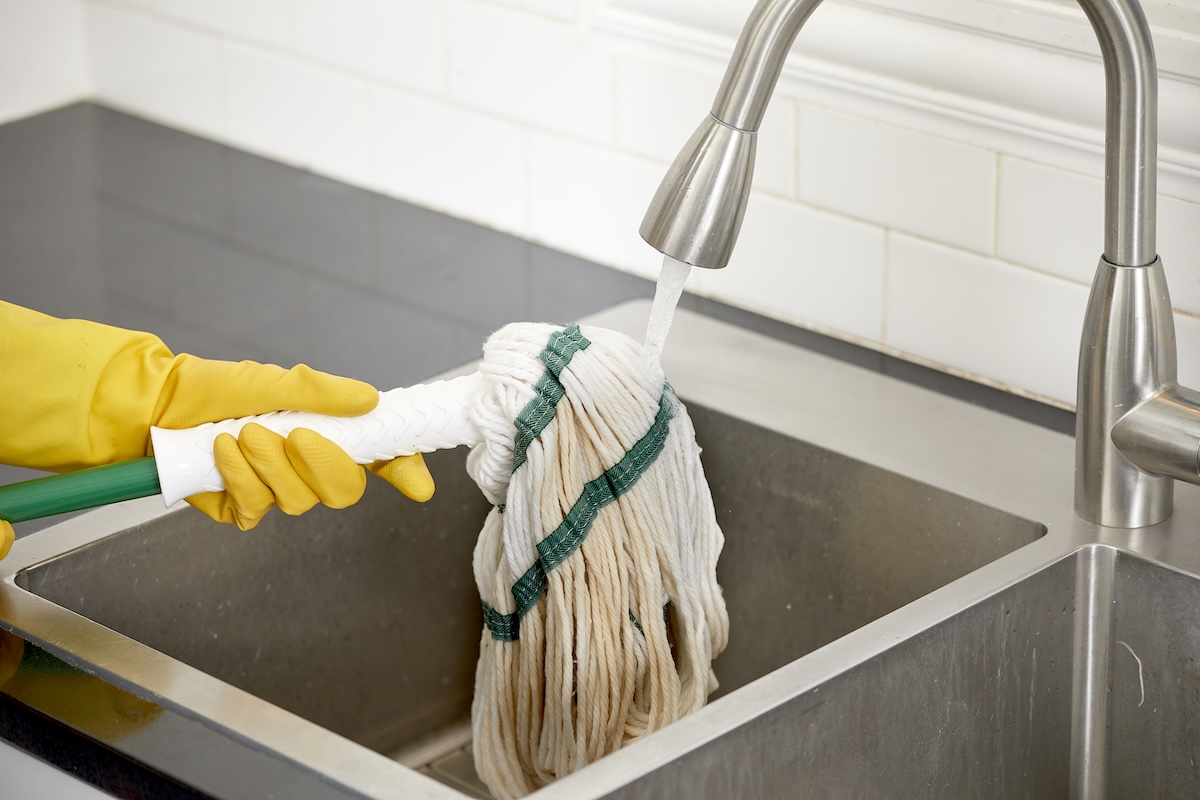 Woman wearing yellow rubber gloves rinses a mop under a running faucet.