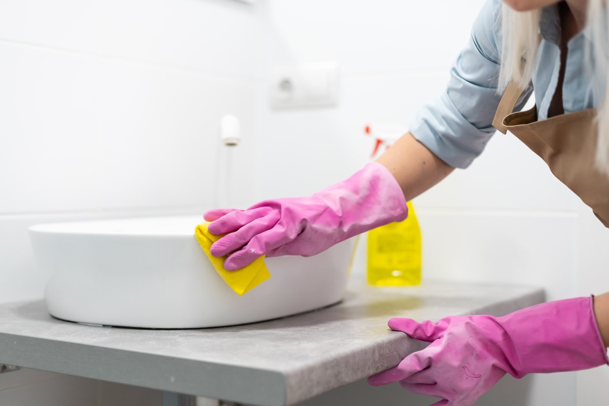 A woman in gloves scrubs a bathroom sink.