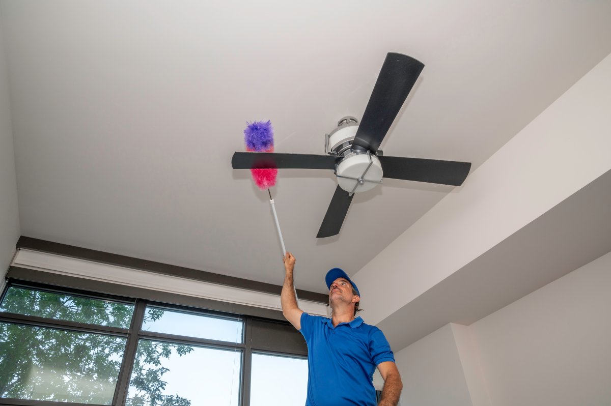 Man cleans dusty ceiling fan blades inside of a home.