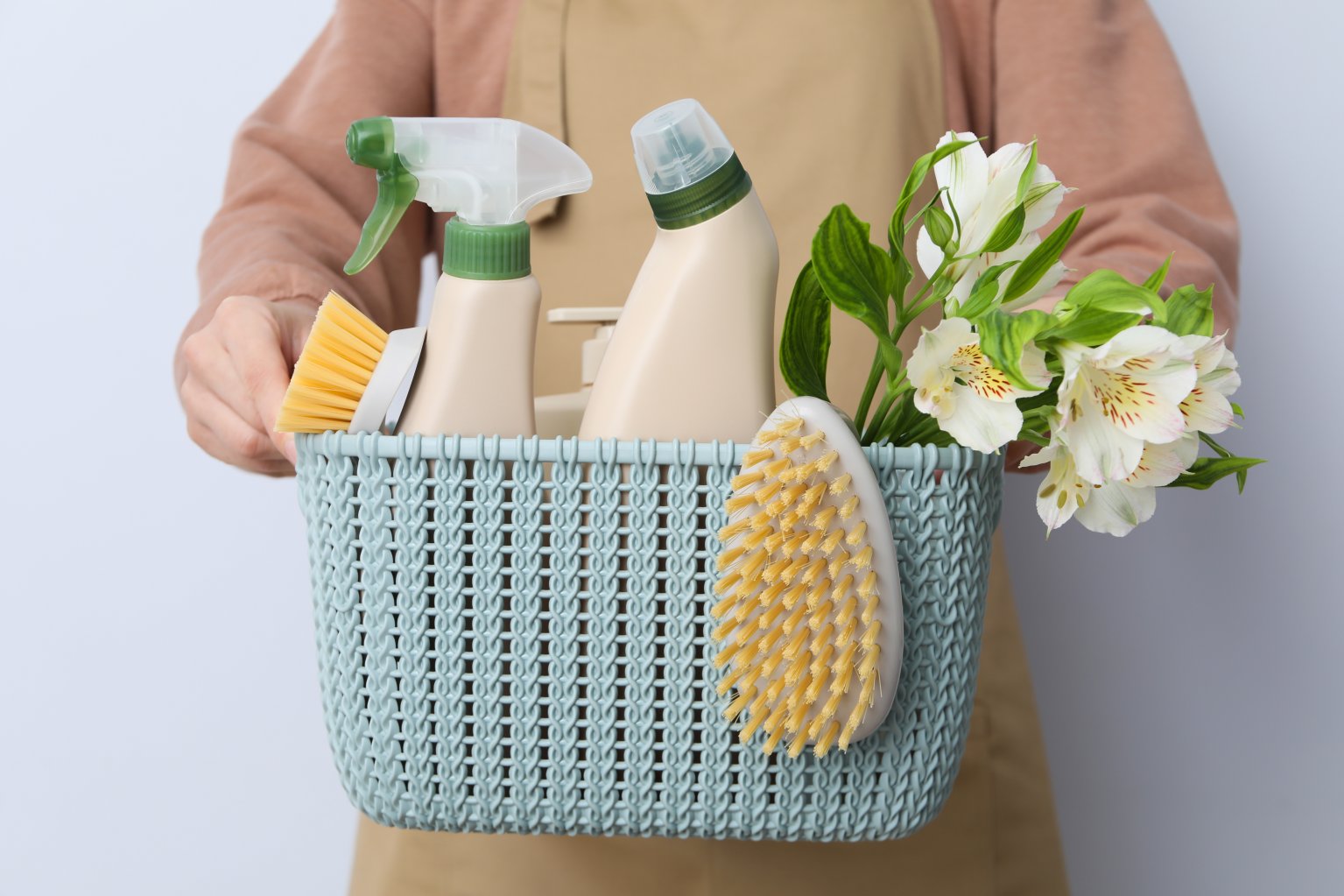 Person in apron holding basket of spring cleaning supplies along with flowers.