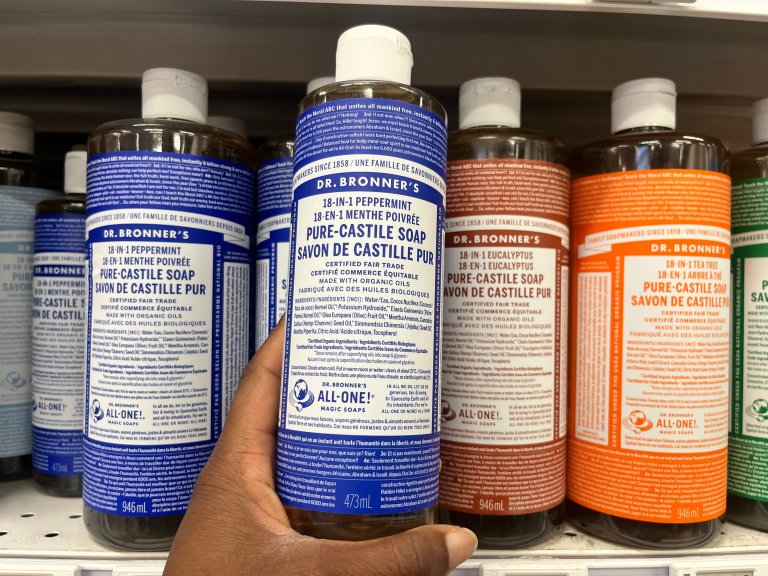 Hand holding a bottle of Dr. Bronner's 18-in-1 peppermint pure castile liquid soap on display on a store shelf.