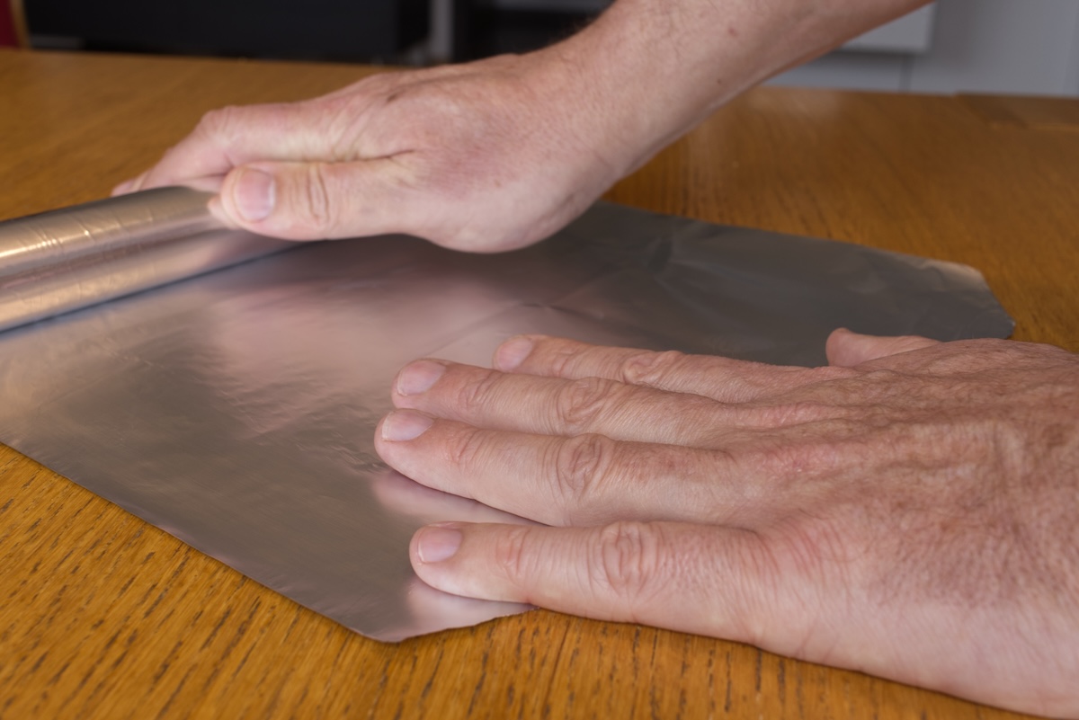 Male hands unroll aluminum foil on a wooden countertop.