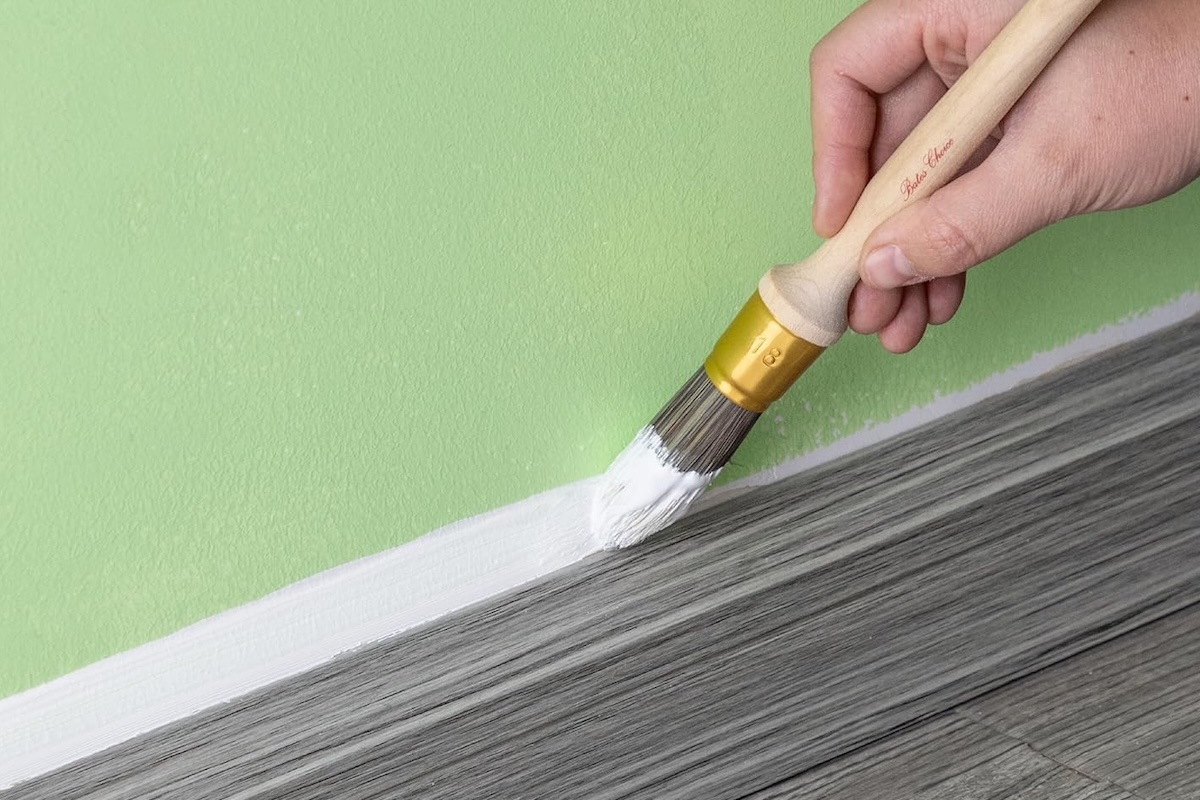 Person uses a Bates round-tipped paintbrush to paint white baseboard trim next to a mint green wall.