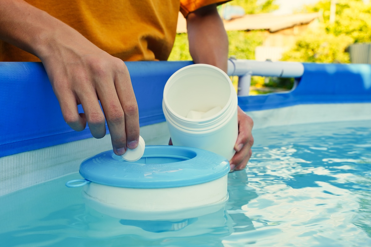 Man's hand holding a white chlorine tablet over swimming pool skimmer.