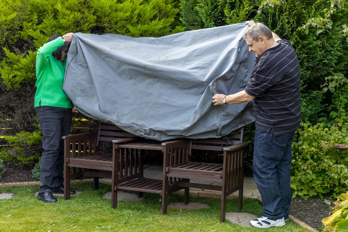 Senior couple puts cover onto outdoor furniture bench set in home backyard.