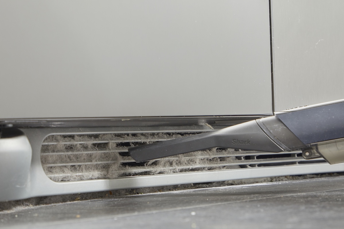 A vaccum attachment cleans dusty coils at the bottom of a refrigerator.