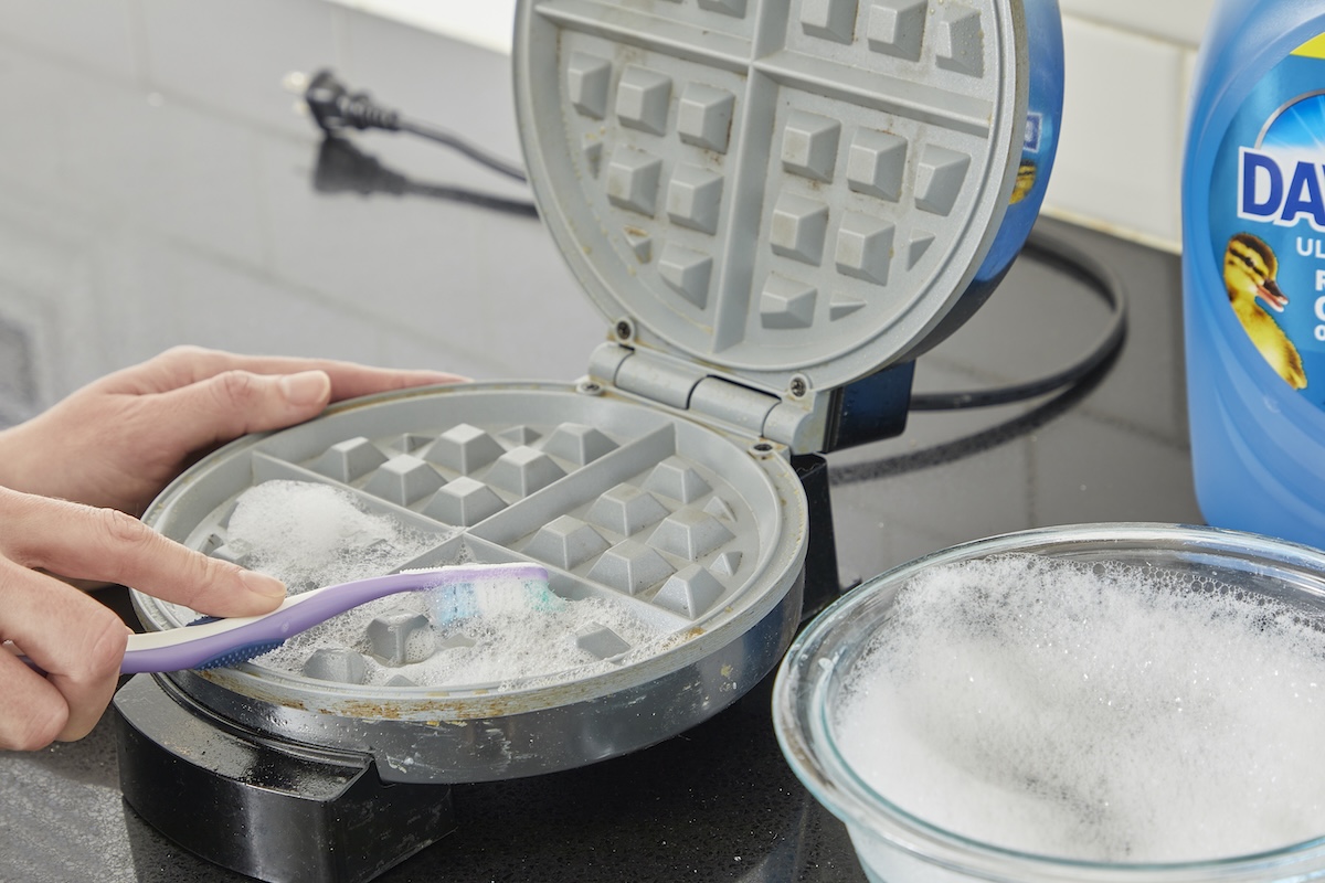 Woman uses a toothbrush and soapy water to scrub a waffle maker.