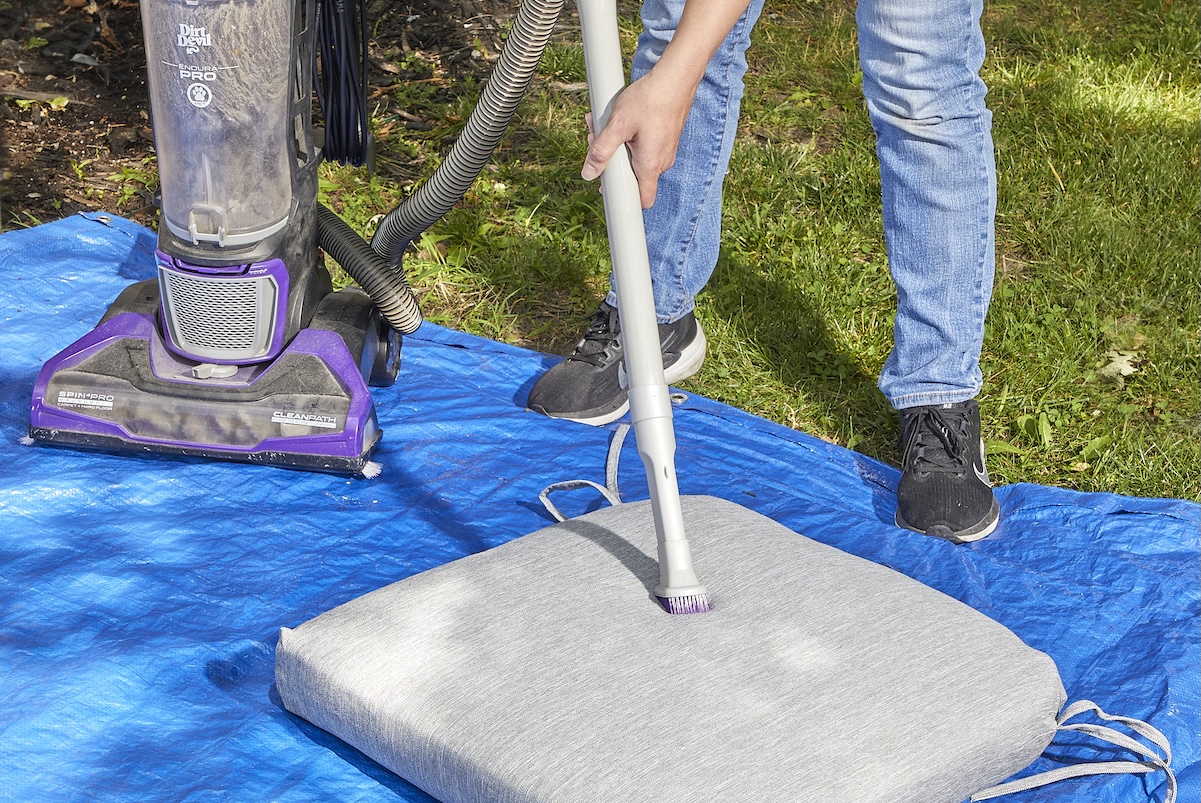 Woman wearing jeans uses hose attachment to vacuum cushion for patio chair, atop a blue tarp outdoors.
